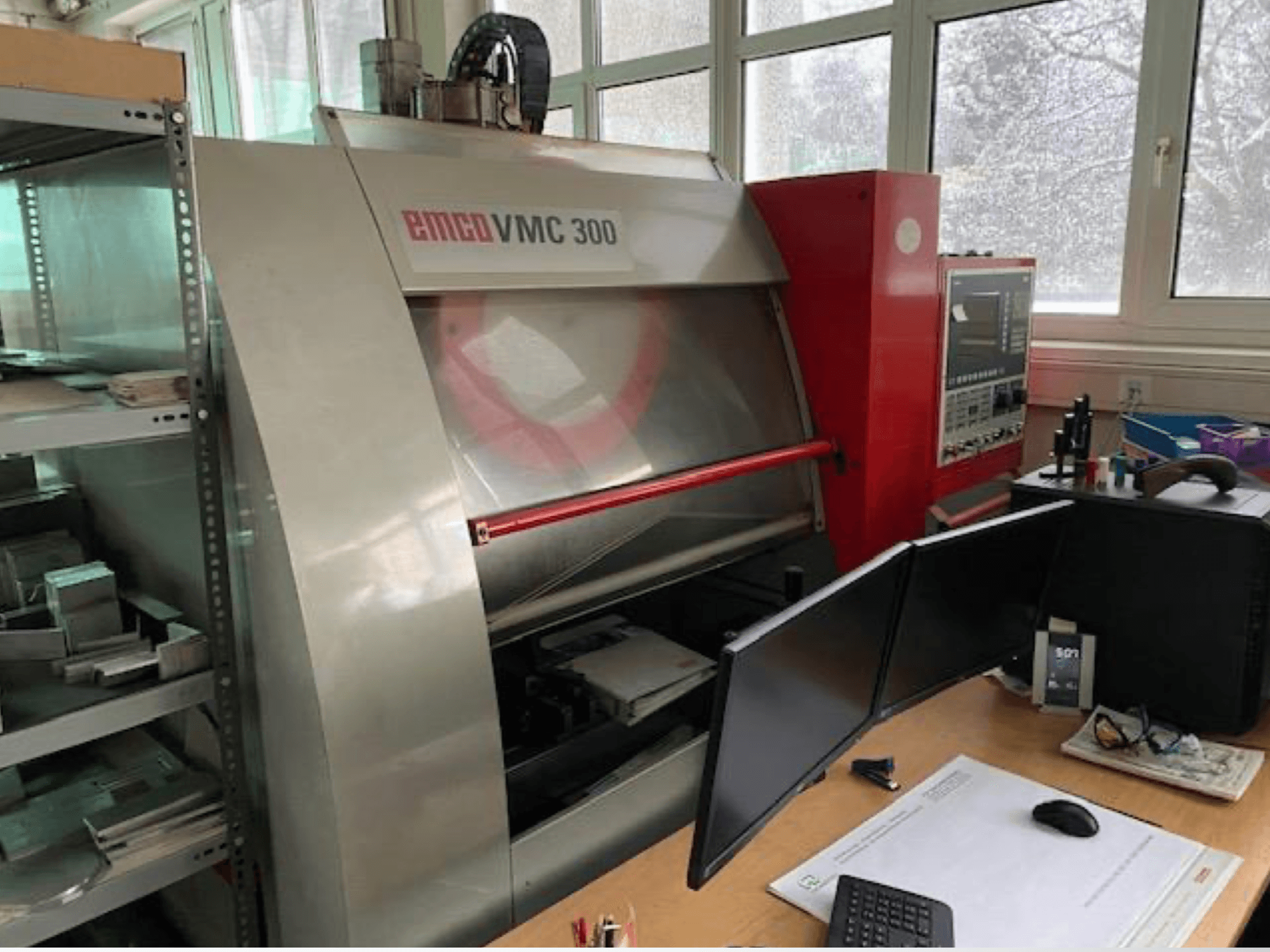 Centro de mecanizado vertical EMCO VMC 300 en un taller; vista lateral en la que se aprecian el panel de control y la estación de trabajo cercana, equipada con dos monitores.