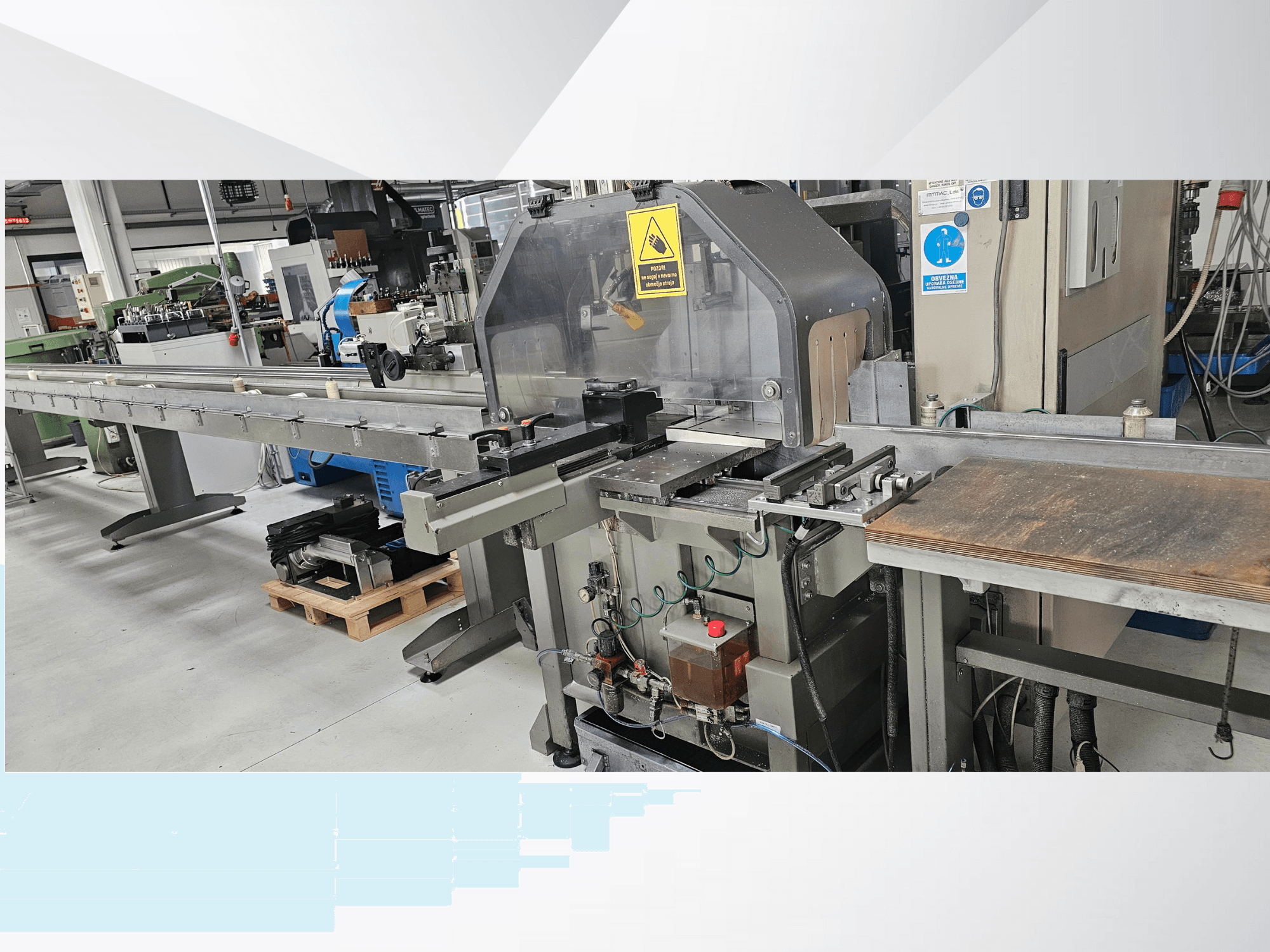 Máquina de corte industrial desde una vista lateral, con un largo transportador, panel de control y advertencias de seguridad.