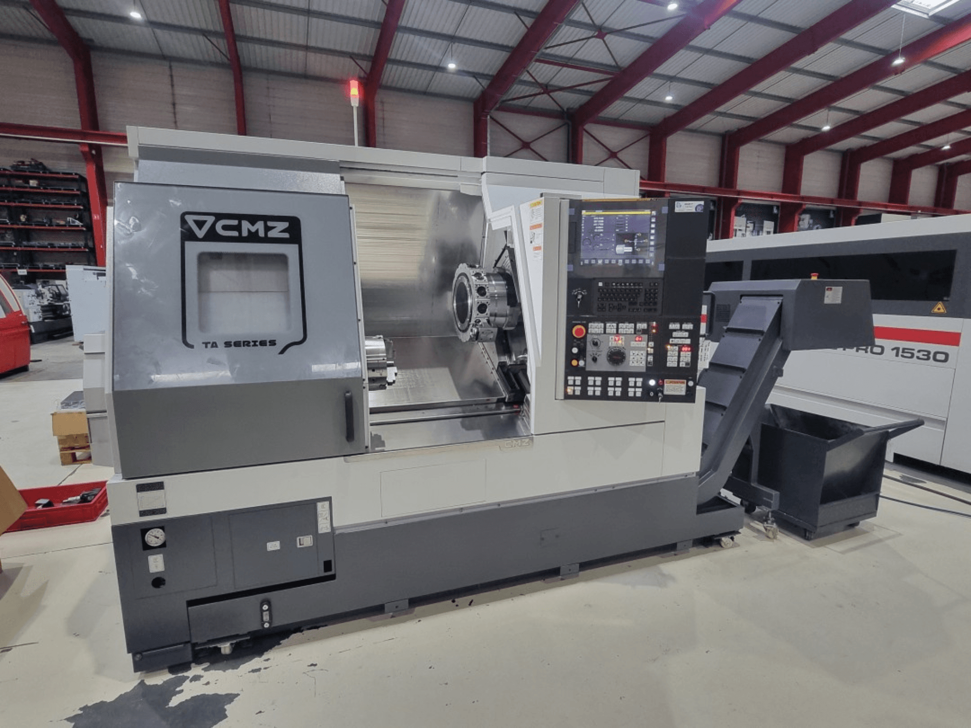 Torno CNC de la serie CMZ TA, vista lateral, con panel de control, área de trabajo y depósito de virutas en un taller.