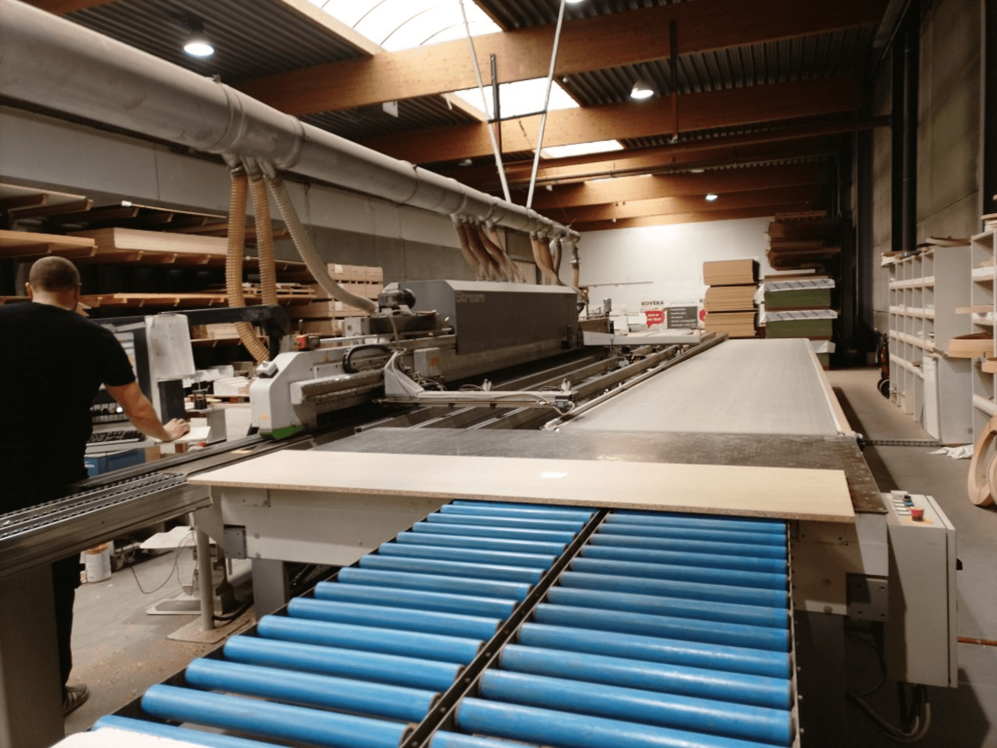 Chapadora de cantos BIESSE Edge Stream B1, vista lateral mostrando el operario, los rodillos azules y el panel de madera sobre el transportador.