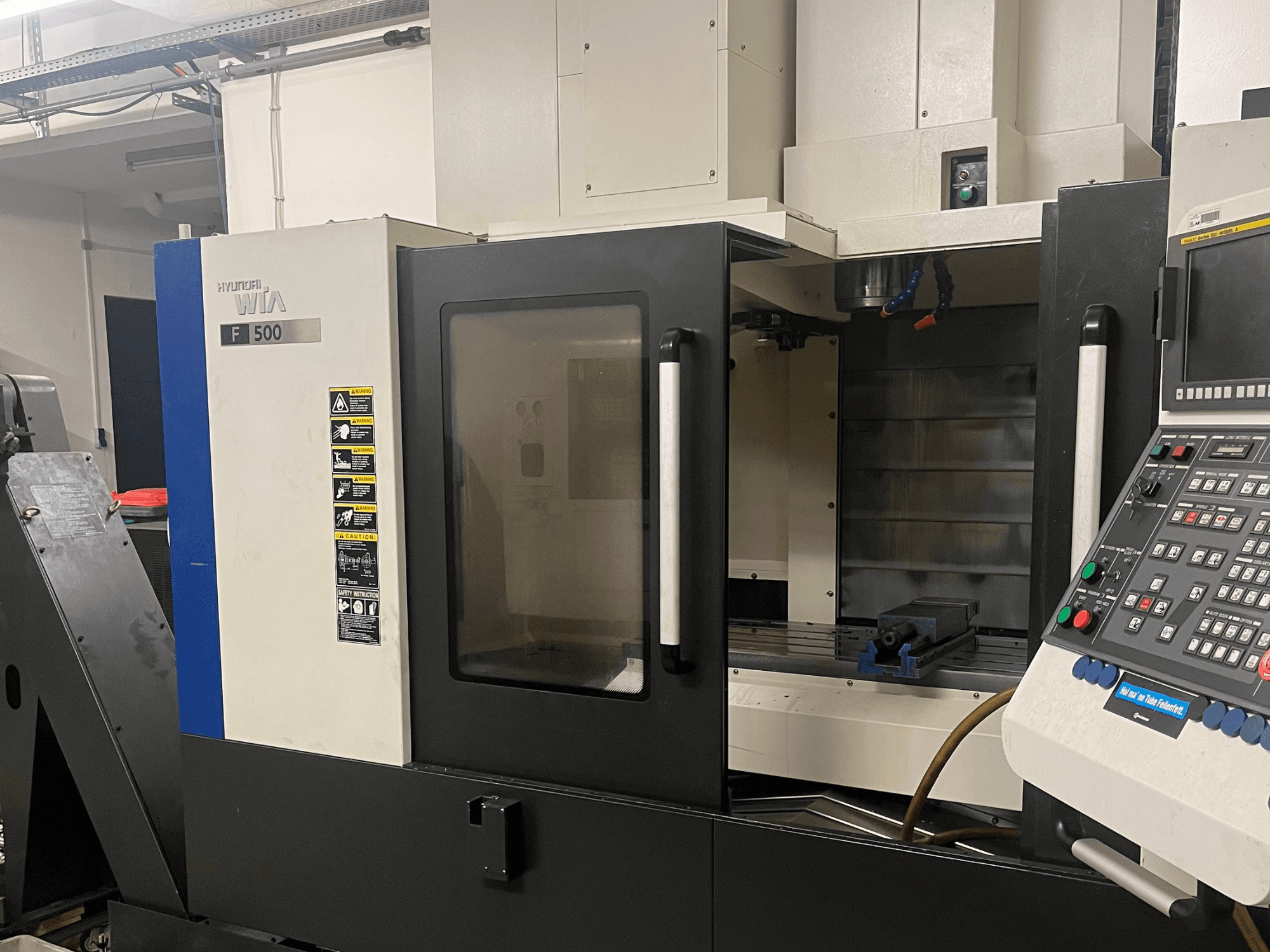 Centro de mecanizado vertical Hyundai Wia F500, vista frontal, en la que se aprecian el panel de control, la zona de trabajo con un tornillo de banco azul y las etiquetas de advertencia.