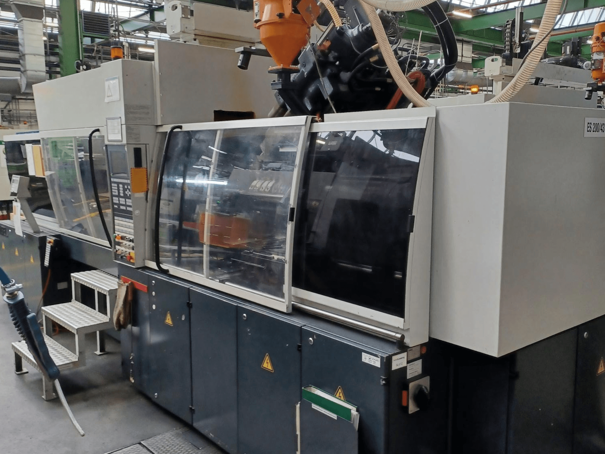 Vista frontal de una máquina de moldeo por inyección de plástico, marca y modelo desconocidos, con puertas de seguridad transparentes y panel de control.