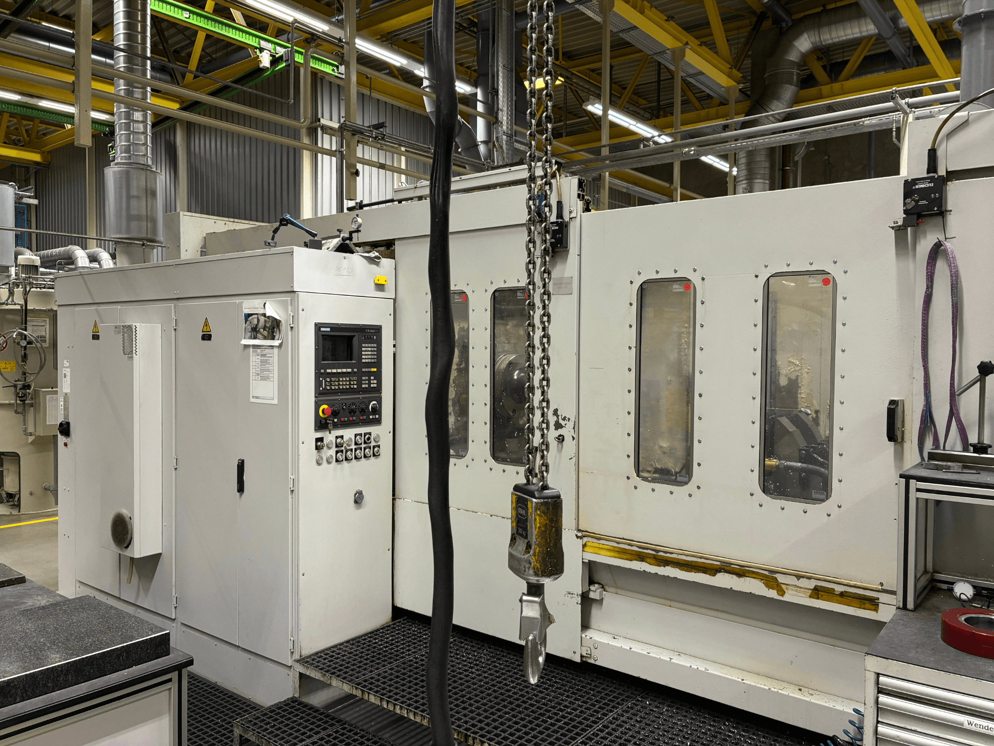 Máquina CNC industrial con panel de control, vista lateral; presenta ventanas, cadenas y componentes metálicos en un entorno de fábrica.