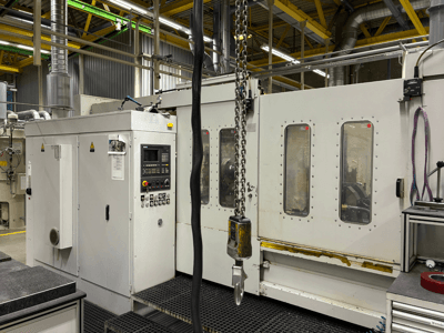 Máquina CNC industrial con panel de control, vista lateral; presenta ventanas, cadenas y componentes metálicos en un entorno de fábrica.